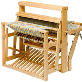 The Schacht 36" High Castle Standard Floor Loom by Schacht Spindle Co. is displayed, featuring a multicolored striped warp. This sturdy wooden loom includes essential components for fabric weaving such as heddles, a reed, and treadles. The structure is supported by a robust wooden base with slatted support.