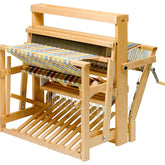 The Schacht 45" High Castle Standard Floor Loom by Schacht Spindle Co. is shown with a partially woven multicolored textile. The yarn threads are neatly arranged, and the loom features various components including a beater, shuttle, heddles, and a hand-crank lever.