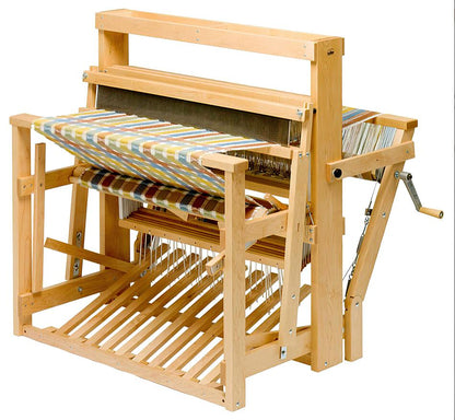 The Schacht 45" High Castle Standard Floor Loom by Schacht Spindle Co. is shown with a partially woven multicolored textile. The yarn threads are neatly arranged, and the loom features various components including a beater, shuttle, heddles, and a hand-crank lever.