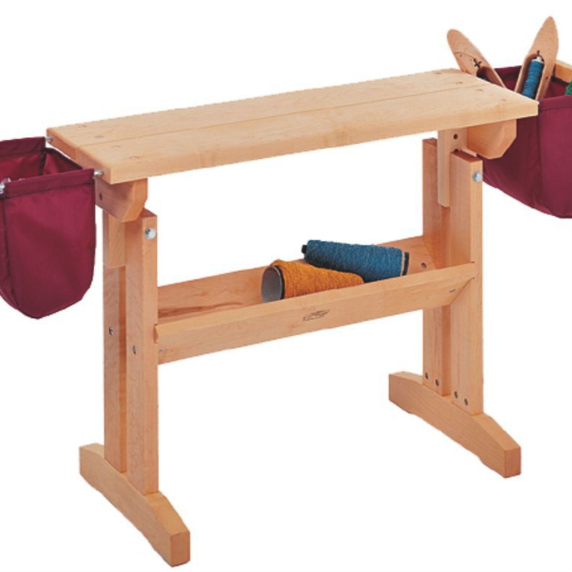 A Schacht Loom Bench by Schacht Spindle Co. with a natural finish features two maroon fabric pouches attached to each end. There is a lower shelf holding blue and orange spools of thread, along with wooden shoe forms visible in the maroon pouches, all set against a white background.