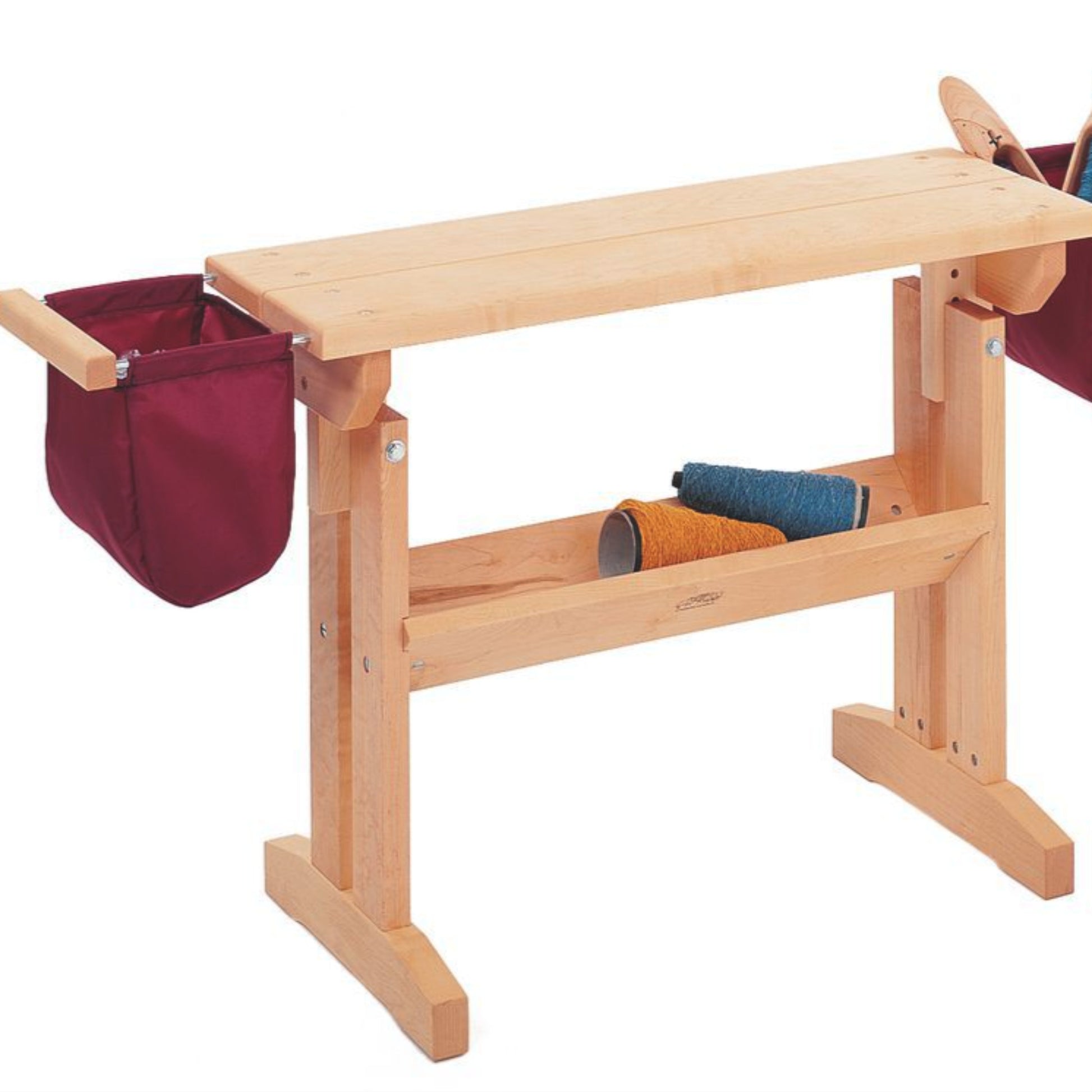 The Schacht Floor Loom Bench Bag by Schacht Spindle Co. incorporates a rectangular tabletop wooden weaving loom, with two crimson side bags attached to each end. It features a lower shelf storing two spools of yarn, one turquoise and one amber. This loom is constructed with sturdy legs and cross beams for enhanced stability.