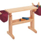 The Schacht Floor Loom Bench Bag by Schacht Spindle Co. is a wooden weaving bench featuring two maroon storage pouches on either side, filled with various weaving tools and materials. There is yarn stored on a lower shelf. The bench has a simple and sturdy design made of light-colored wood.