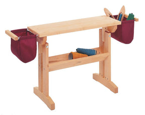 The Schacht Floor Loom Bench Bag by Schacht Spindle Co. is a wooden weaving bench featuring two maroon storage pouches on either side, filled with various weaving tools and materials. There is yarn stored on a lower shelf. The bench has a simple and sturdy design made of light-colored wood.