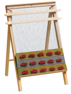 The Schacht School Loom by Schacht Spindle Co. stands upright with a partially completed textile on it. The textile features a pattern of red floral motifs on an olive green background, bordered by narrow black and white stripes, with a gray section between green areas. A weaving needle rests nearby, ready for use.