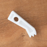 A Table Loom & Rigid Heddle Ratchet Dog from Schacht Spindle Co., crafted in the shape of a traditional key, rests on a wooden surface. This white plastic tool boasts a grooved edge and includes a hole near the top, making it ideal for precisely adjusting Schacht table looms.