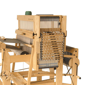 The Louët Megado Mechanical Dobby Head by Louët Inc. features a wooden frame with metal heddles and threads meticulously set up for weaving. The central area displays warp threads secured tightly, and the structure includes various components such as harnesses, a reed, and a cloth beam. A green spool of thread is placed on top.