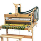 The Louët Magic Dobby 24-shaft Floor Loom without Dobby Control Head by Louët Inc. showcases yellow and black threads partially woven into fabric. This sophisticated wooden loom is equipped with a seat, an array of pulleys, and levers, including an intricate mechanism on the right side. Various tools and materials can be seen on the loom and surrounding surfaces.