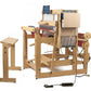 A Louët Megado Floor Loom without Dobby Control Head by Louët Inc. with various mechanical components and threads is set up in a workspace. A stool is positioned to the side, and the loom features a digital display screen. An electrical cord is connected to the loom, implying it is electronically operated.