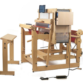 A Louët Megado Floor Loom without Dobby Control Head by Louët Inc. with various mechanical components and threads is set up in a workspace. A stool is positioned to the side, and the loom features a digital display screen. An electrical cord is connected to the loom, implying it is electronically operated.