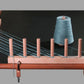 The Louët Warping Posts, crafted from premium beech hardwood, are securely clamped to a table. Blue yarn winds meticulously around its parallel pegs and leads to a large spool on the right. This efficient warping solution is perfect for measuring and preparing yarn for weaving, making it especially useful for rigid heddle weavers.