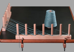 The Louët Warping Posts, crafted from premium beech hardwood, are securely clamped to a table. Blue yarn winds meticulously around its parallel pegs and leads to a large spool on the right. This efficient warping solution is perfect for measuring and preparing yarn for weaving, making it especially useful for rigid heddle weavers.