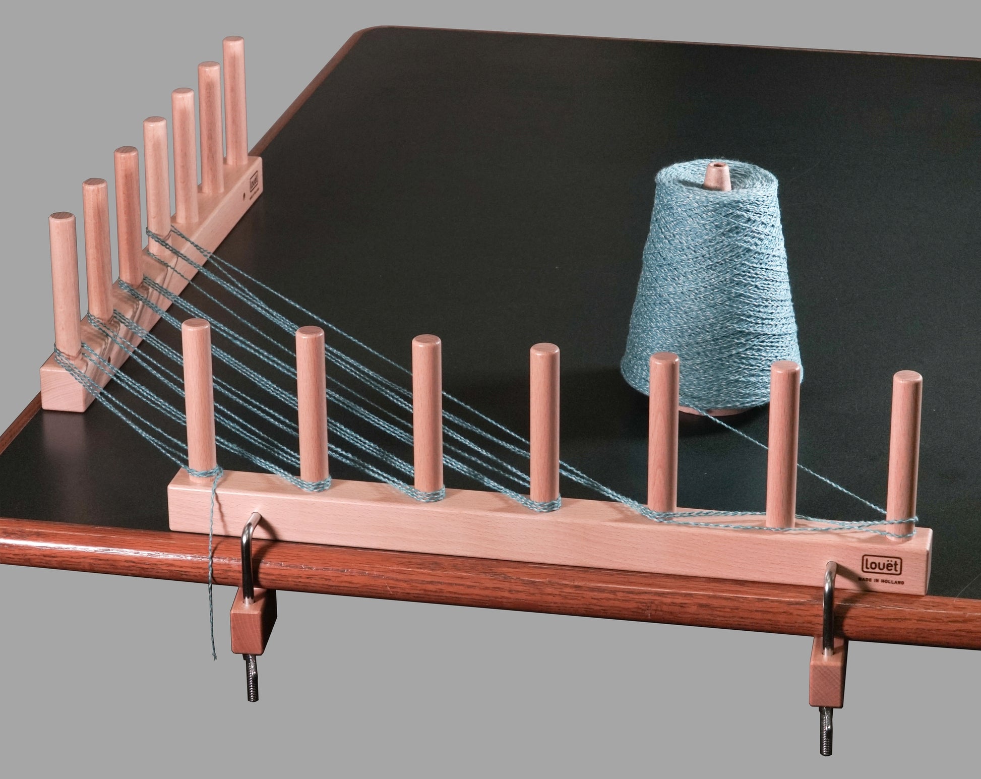 The Louët Warping Posts from Louët Inc., crafted from beech hardwood with multiple pegs, are securely attached to the edge of a table. Several strands of blue yarn are threaded through the pegs, and a large spool of the same blue yarn rests on the table next to this ideal warping solution for rigid heddle weavers.