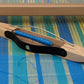 A Louët Flying Dutchman Shuttle, 34 cm (13.4"), rests on a blue and yellow loom. The wooden shuttle from Louët Inc. has a black handle and is threaded with blue yarn. The vibrant woven fabric beneath features stripes and a multicolored pattern.