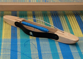 A Louët Flying Dutchman Shuttle, 34 cm (13.4"), rests on a blue and yellow loom. The wooden shuttle from Louët Inc. has a black handle and is threaded with blue yarn. The vibrant woven fabric beneath features stripes and a multicolored pattern.