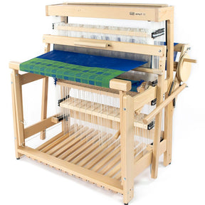 The Louët Inc. Louët Spring 2, 8-shaft Floor Loom is a robust and expertly crafted apparatus featuring multiple heddles and a partially woven fabric in vibrant blue and green hues. This sophisticated loom includes various levers and pedals, with an array of threads meticulously threaded throughout the frame. The overall setup is built with sturdy wooden frames and beams for enhanced durability.