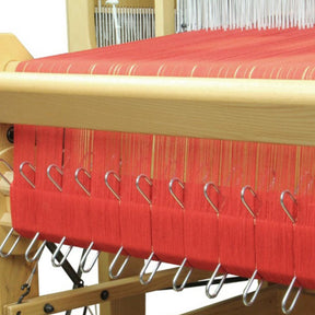 A Louët Inc. loom equipped with the Louët Sectional Warp Kit for the Floor Looms, featuring tightly strung red threads in 1" or 2 cm sections, held by multiple metal hooks. The loom’s sectional warp beam and various mechanisms are partially visible, indicating an ongoing weaving process.