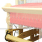 A close-up view of the Louët Second Warp Beam for the Megado Loom from Louët Inc., showcasing pink threads being meticulously woven. The loom's various components, such as metal hooks and gears, are clearly visible. This intricate setup highlights the detailed process of weaving fabric.