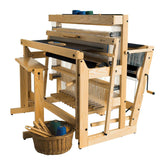 The Louët Delta Floor Loom by Louët Inc., made of wood with a complex arrangement of threads and mechanical parts, is accompanied by a basket of colorful yarns and weaving tools on the floor near it, while the top holds additional spools of yarn, making it an exercitation ullamco masterpiece for any artisan.