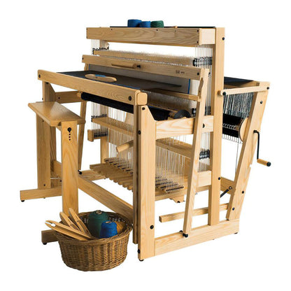 A large Louët Delta Floor Loom designed for weaving textiles sits prominently, featuring various spools, threads, and a side lever for adjustment. A basket holding spools of colored yarn and some wooden tools is placed beside it. The structure is primarily light-colored wood by Louët Inc., making exercitation ullamco an elegant choice.
