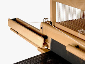 Close-up of the Louët Fly Shuttle Device/Mechanism for Floor Looms by Louët Inc., with a shuttle containing white thread positioned on the left side. Multiple white threads run vertically through the comb-like reed, while partially woven black fabric is visible on the bottom section of the loom, showcasing intricate weaving.