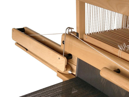 A detailed view of a wooden weaving loom in action, featuring the Louët Fly Shuttle Device/Mechanism for Floor Looms by Louët Inc., with heddles and black threads being woven. The loom components are crafted from light-colored wood, and white strings are arranged across the heddles.