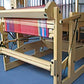 A beige wooden Megado loom by Louët Inc. with multicolored threads arranged for weaving, featuring the Louët Second Warp Beam. The structure is set up in a well-lit room, showcasing intricate patterns being woven onto the fabric. The background is slightly blurred, focusing on the loom's detailed design and second beam.