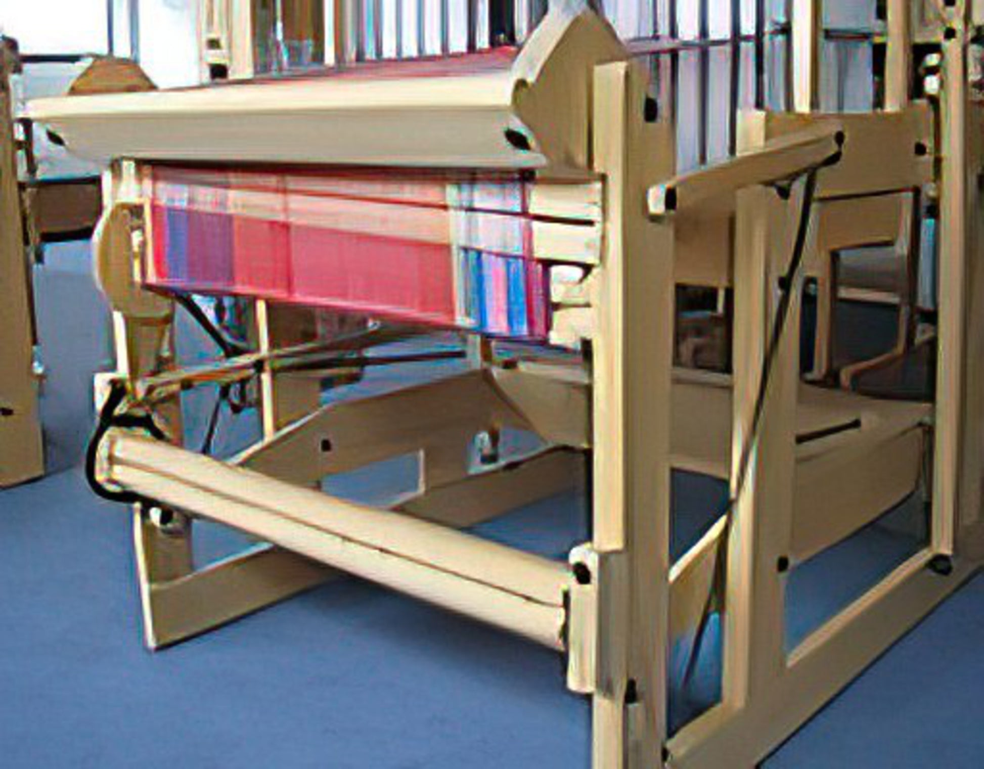 A beige wooden Megado loom by Louët Inc. with multicolored threads arranged for weaving, featuring the Louët Second Warp Beam. The structure is set up in a well-lit room, showcasing intricate patterns being woven onto the fabric. The background is slightly blurred, focusing on the loom's detailed design and second beam.