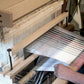 A person is skillfully weaving fabric with parallel stripes in various shades of black, white, and red on a wooden David I loom. Their hands are clearly visible as they manipulate the threads using the Louët Sliding Beater for David Looms by Louët Inc. The hand-operated loom, complete with its overhead beater, is set up indoors.