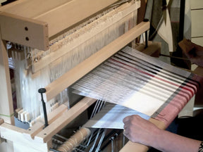 A person is skillfully weaving fabric with parallel stripes in various shades of black, white, and red on a wooden David I loom. Their hands are clearly visible as they manipulate the threads using the Louët Sliding Beater for David Looms by Louët Inc. The hand-operated loom, complete with its overhead beater, is set up indoors.