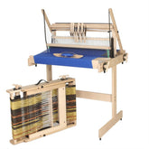 Image of a wooden Louët Jane Table Loom by Louët Inc. for weaving textiles. The loom features a partially woven blue fabric on its frame and an attached spool of threads in various colors (yellow, black, brown). It stands on the floor with an adjustable height and sturdy frame, ideal for your next weaving project.