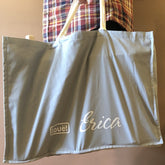 A person wearing a plaid shirt holds the Louët Erica Cotton Carrying Bag by Louët Inc., a light blue tote with cream-colored handles. The bag features white embroidered lettering, with "Erica Loom" in cursive and "Lovéit" in a logo style.