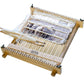 The Louët Lisa Tapestry Loom by Louët Inc. is a small tabletop wooden loom featuring white threads and partially woven fabric. The rectangular frame contains metal pins along the top and bottom edges to securely hold the threads in place, and some yarn is visible being woven through it, indicating that the loom is currently in use.