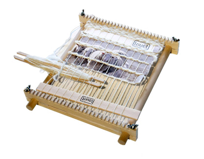 A Louët Lisa Tapestry Loom by Louët Inc., featuring a wooden frame with white threads arranged vertically and a shuttle resting on top. Some fabric is being woven in the middle, showcasing the loom's functionality. The simple rectangular frame has adjustable parts, making it perfect for intricate weaving projects.