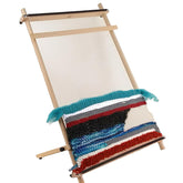 A Louët Lisa Tapestry Loom by Louët Inc. with partially woven fabric in various colors including red, blue, black, and white. The woven section displays horizontal patterns and textures, with some loose threads hanging. The loom stands against a white background.