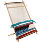 A Louët Lisa Tapestry Loom by Louët Inc. with partially woven fabric in various colors including red, blue, black, and white. The woven section displays horizontal patterns and textures, with some loose threads hanging. The loom stands against a white background.