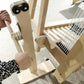 An individual wearing a black and white patterned shirt is using a Louët Loom Lift Device for Spring 2 from Louët Inc., turning its wooden crank handle. White threads are stretched across the device, showing it in use for weaving or spinning yarn, similar to a traditional loom, against a beige carpeted floor.