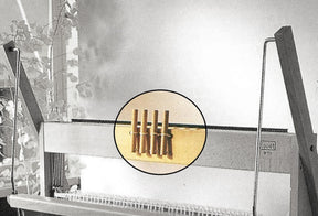 A Louet Inc. table loom with a magnified view highlights a set of four wooden pegs or knitting tools indoors near a window with visible potted plants in the background. The magnified view draws attention to specific components, including the Louët Table Loom Toggle Handle replacement part.