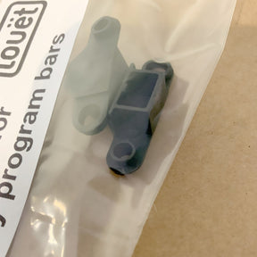 A small black plastic component is sealed in a clear plastic bag. Next to it, there is a white label with partially visible text, including the words "dobby bars." The background features a light brown surface, suggesting it's meant to replace non-functional shaft parts. This is the Louët Coupling piece for dobby bars (2 pack) from Louët Inc.