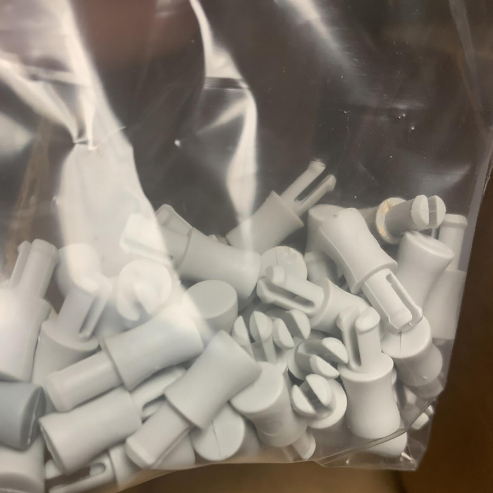 A sealed, clear plastic bag from Louët Inc. contains multiple small, gray cylindrical pieces with varying connectors and attachments at the ends. These objects are closely packed together and are part of the "Louët Pegs for Dobby Bars (set of 50).