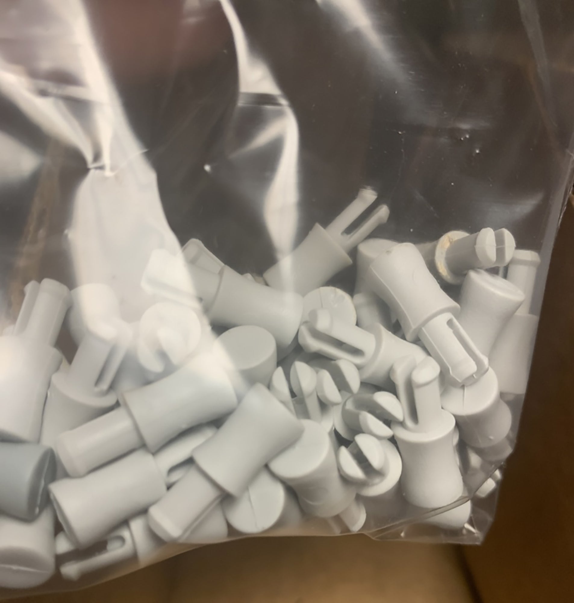 A clear plastic bag filled with numerous small white plastic components of various shapes and sizes, likely Louët Pegs for Dobby Bars (set of 50) from Louët Inc. The bag is slightly crumpled, and some parts are easily visible through the transparent material.