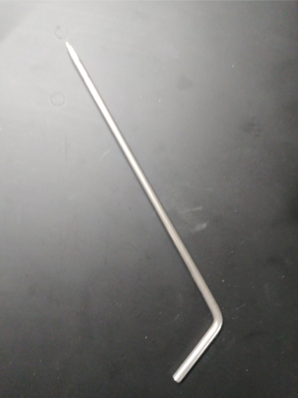 A Louët Weaving Loom Blocking Pin, a silver L-shaped metal rod by Louët Inc., with one end slightly pointed, laying on a dark surface.