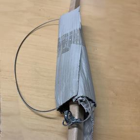 A makeshift clamp constructed from a wooden stick, gray duct tape, the Louët Spring Brake Cable by Louët Inc., and metal attachments on a light wooden surface. The cable forms a loop, indicating that this DIY solution aims to hold or secure items effectively.