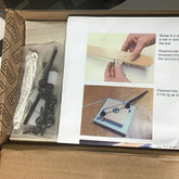 An open box reveals a printed manual with detailed instructions and images of hands-on repair processes. To the left of the manual, several small packaged components, including metal rods and screws, are neatly arranged. The box, lined with paper featuring a repeating "Louët Octado" pattern, hints at the intricacies involved in assembling the Louët Octado Mechanical Dobby Repair Kit by Louët Inc.