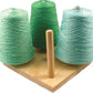 Three large cones of yarn in varying shades of green and blue are displayed on a wooden base with a central peg. This setup, evocative of the Harrisville 4-Peg Cone Holder by Harrisville Designs, is perfect for small studios, offering an economical tool for holding yarn cones neatly wound and ready for knitting or other crafts.