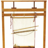 The Navajo Style Loom by Dovetail is a sturdy wooden weaving loom with a partially finished textile. This loom, crafted in the Navajo style, features vertical white warp threads and a horizontal wooden rod that secures them. Several tools and strings are attached to facilitate the weaving process.