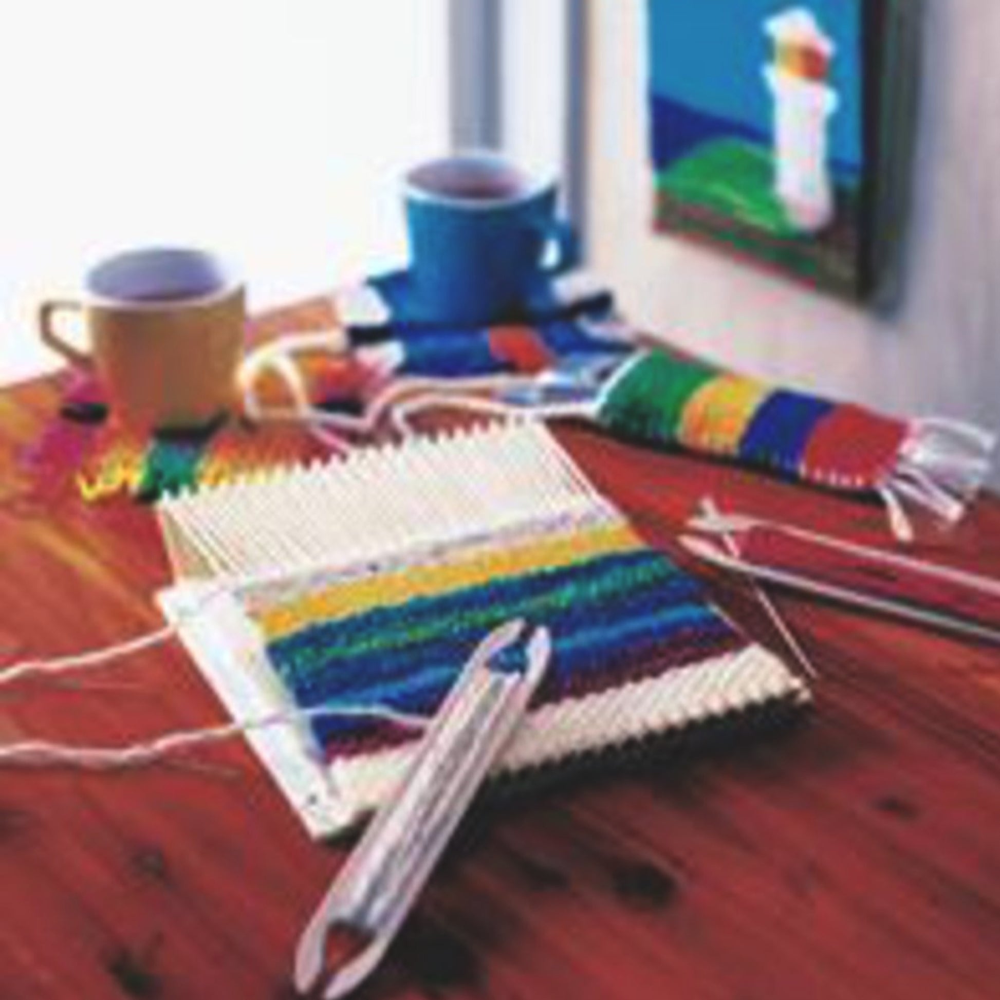 A Schacht Spindle Co.'s Schacht Mini Loom Weaving Kit is set on a wooden table, displaying a colorful, partially woven fabric. Nearby are a blue and yellow cup, small pieces of finished woven cloth, a painting on the wall, and crafting tools scattered around.