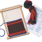 A small Harrisville Designs Peg Loom with a partially completed design in red, green, and yellow yarn. Nearby are a skein of multicolored yarn, a needle with blue thread, a spool of white string, and an instructional booklet titled "Weaving with Your PegLoom." Perfect for kids weaving projects.