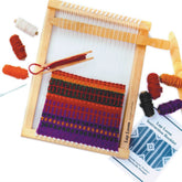 Displayed is a Harrisville Designs Harrisville Peg Loom with Accessories, showcasing an in-progress weaving project adorned with vibrant yarns in purple, orange, and black. Surrounding the hardwood peg loom are spools of yarn in multiple colors, a needle, and a Lap Loom Instruction Booklet, making it an ideal choice for kids' weaving activities.