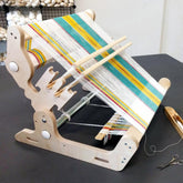 The Glimakra Siru Rigid Heddle Loom w/shuttle and warping peg, crafted from birch plywood, displays a partly woven fabric with white, yellow, green, and blue stripes. The Glimakra loom rests on a dark surface, accompanied by wooden shuttles and scissors. Rolls of yarn are visible in the background, enhancing the ergonomic weaving experience.