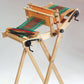 A Glimakra Emilia Rigid Heddle Loom by Glimakra on a foldable stand with a multicolored fabric partially woven. The fabric features red, green, and yellow stripes. The loom includes solid metal gears and handles for adjusting and tightening the threads, accompanied by detailed weaving instructions.